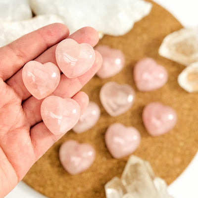 Hand holding 3 Rose Quartz Hearts with others blurred on corkboard