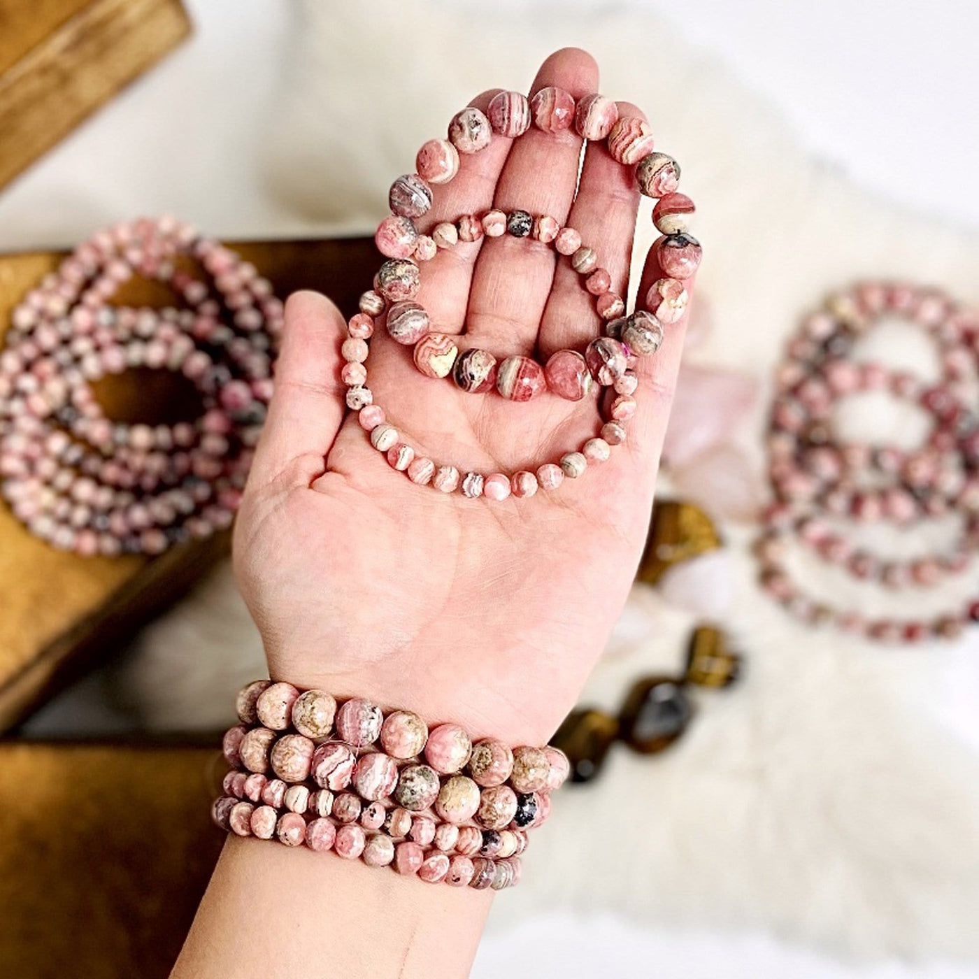 Hand wearing Rhodochrosite Round Bead Bracelets and holding up 2 more bracelets with more blurred in the background