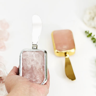 Hand holding up silver Rose Quartz Butter Spreader with gold one blurred in the background
