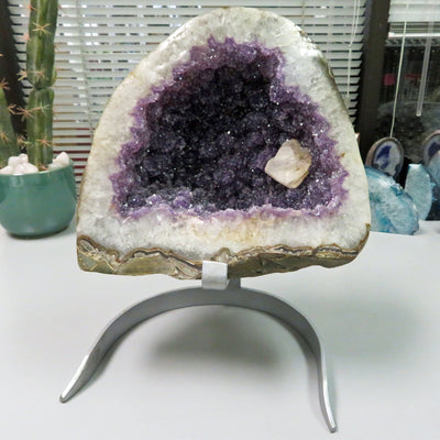 Large amethyst geode formation. It has a white border with amethyst cluster purple inside and one calcite white formation on the right bottom side. It is on a silver metal stand.