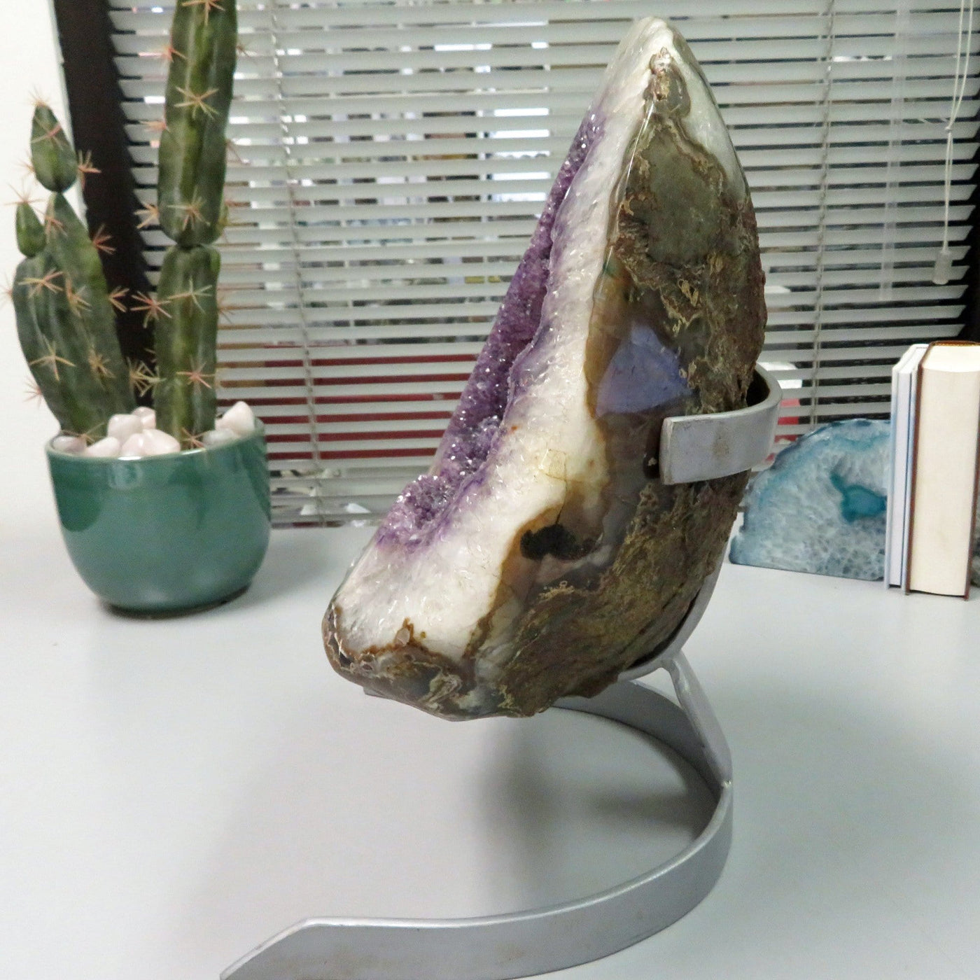Side view of Large amethyst geode formation. It has a white border with amethyst cluster purple inside and one calcite white formation on the right bottom side. It is on a silver metal stand.