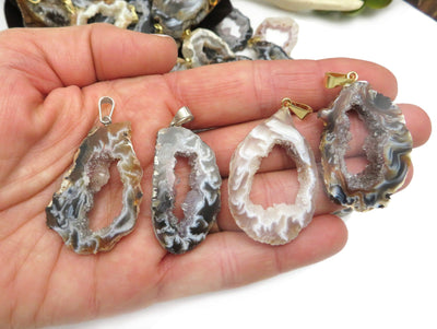 Four agate geode druzy pendants shown in a hand, two with silver bails and two with gold bails. Multiple pendants in the background.