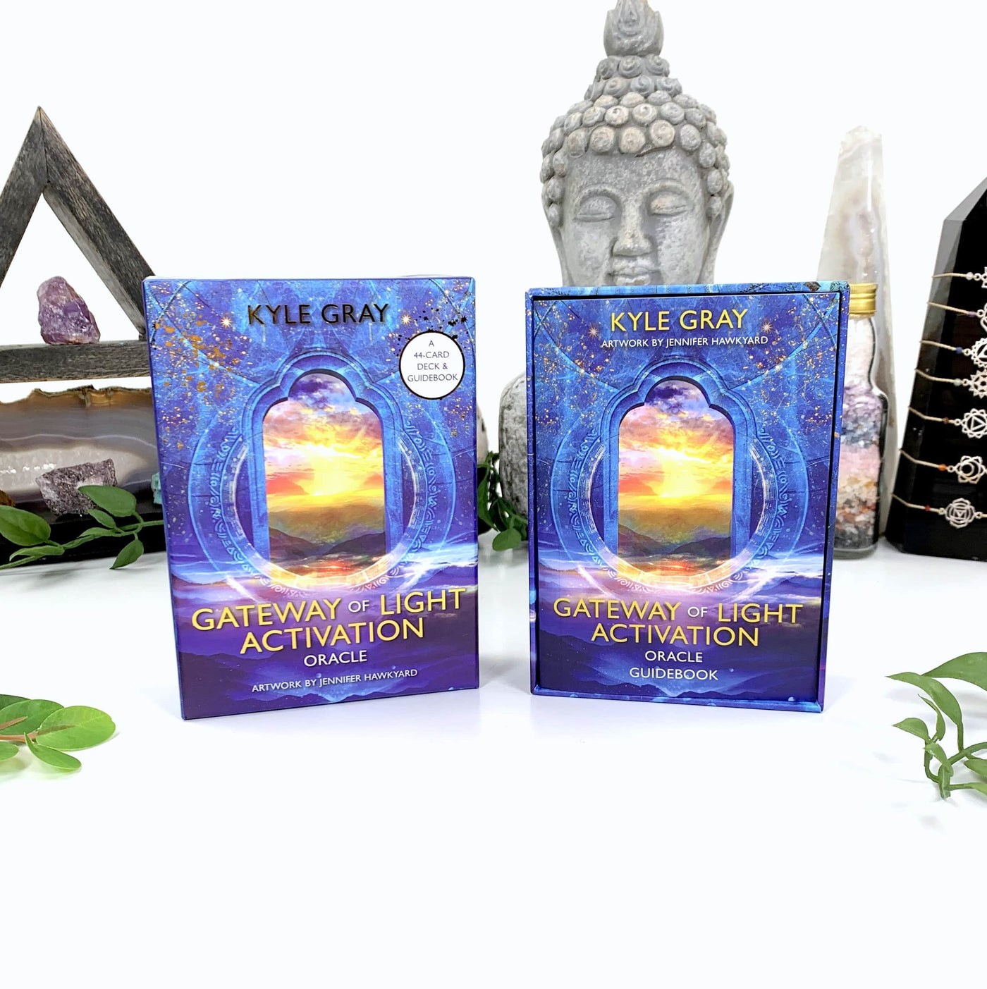 Image of back and front of the oracle card box on a white background with a budha statue as a background prop.  Box is blue with a printed window showing a sunset over mountains.