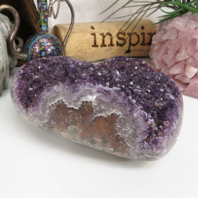Side view of Large amethyst cluster heart on a white background with other crystals and decorations around it.