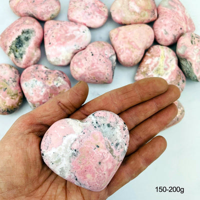 Hand holding up 150-200g Rhodonite Heart with others blurred in the background