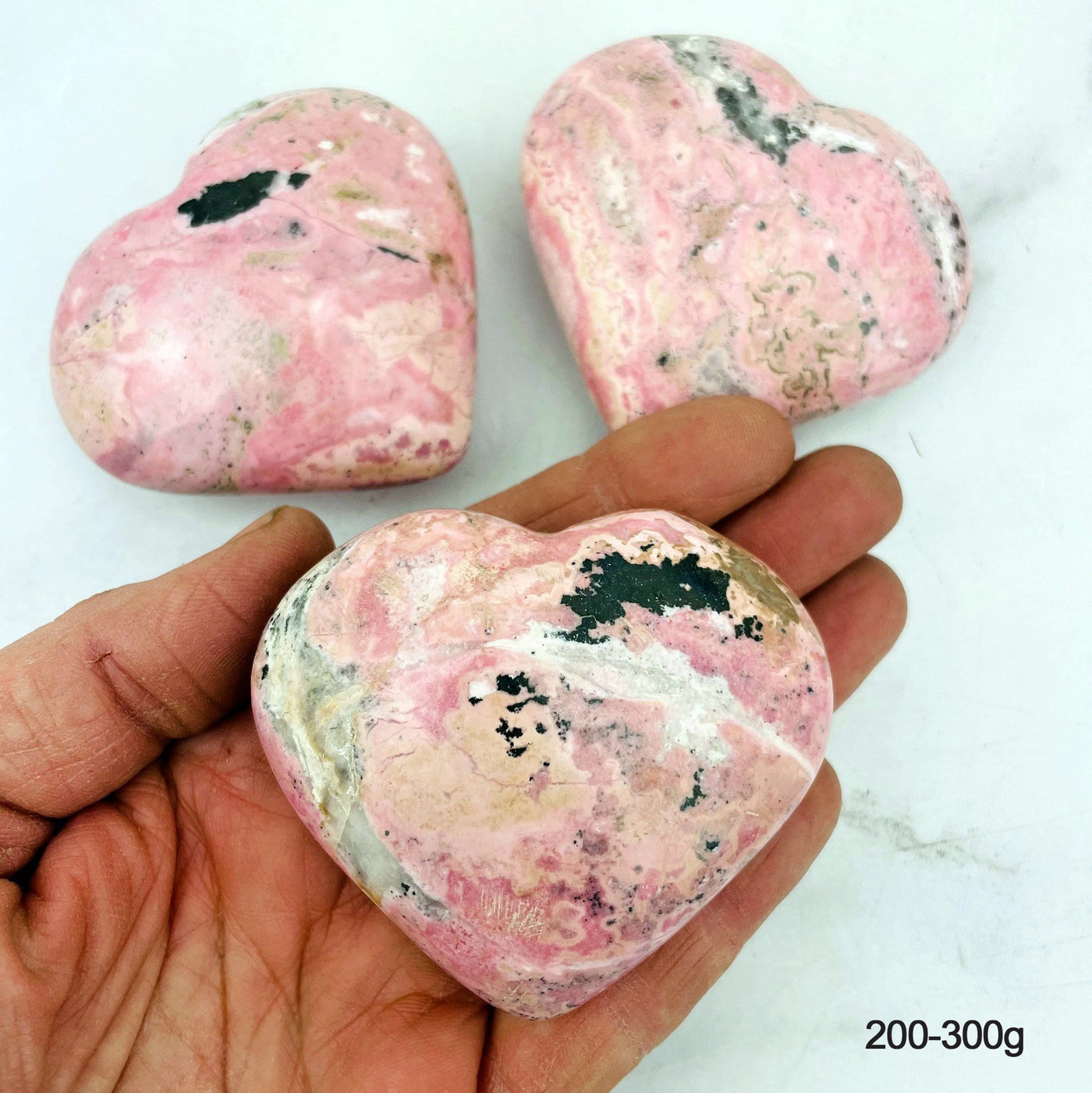 Hand holding up 200-300g Rhodonite Heart with 2 others in the background