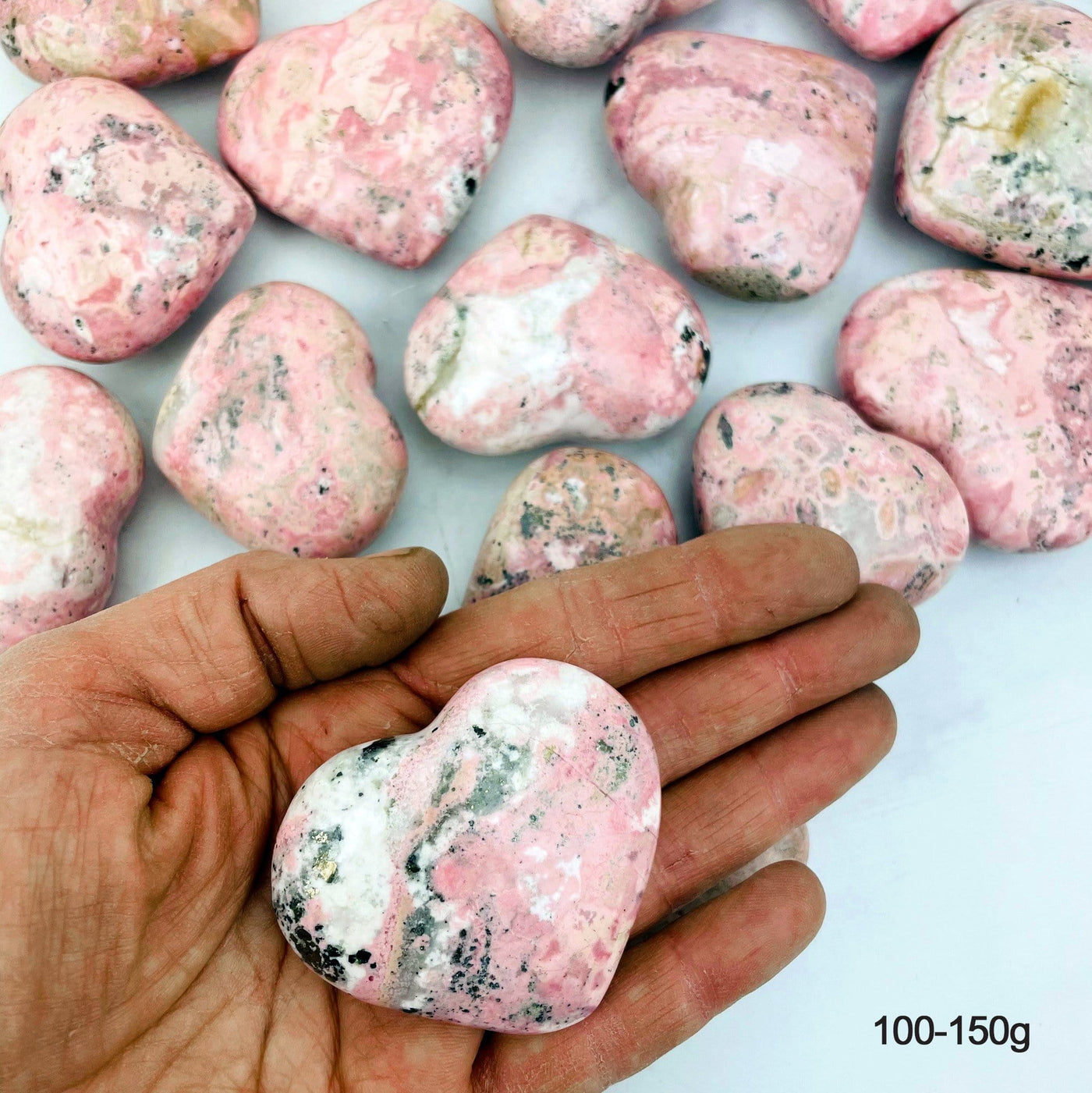 Hand holding up 100-150g Rhodonite Heart with others scattered in the background