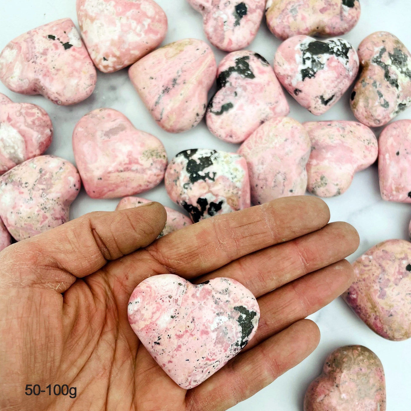 Hand holding up 50-100g Rhodonite Heart with others scattered in the background