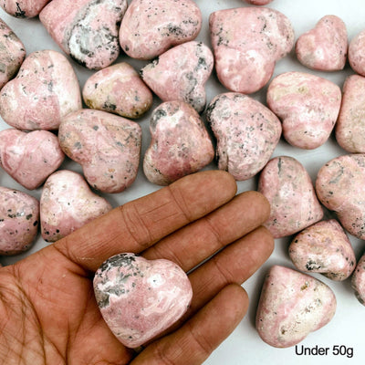 Hand holding up Rhodonite Heart under 50g with others scattered in the background
