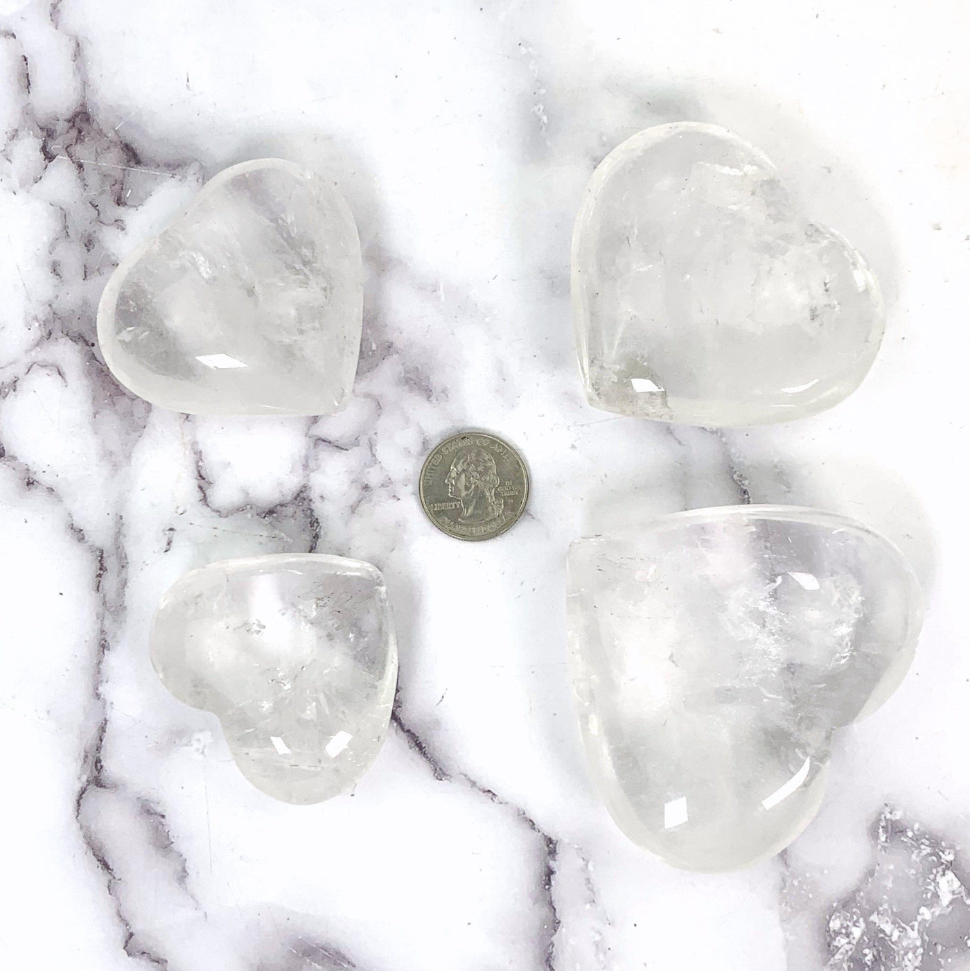 Crystal Quartz Heart--Top view of all 4 hearts comparing a quarter for size reference, on a white marble background.