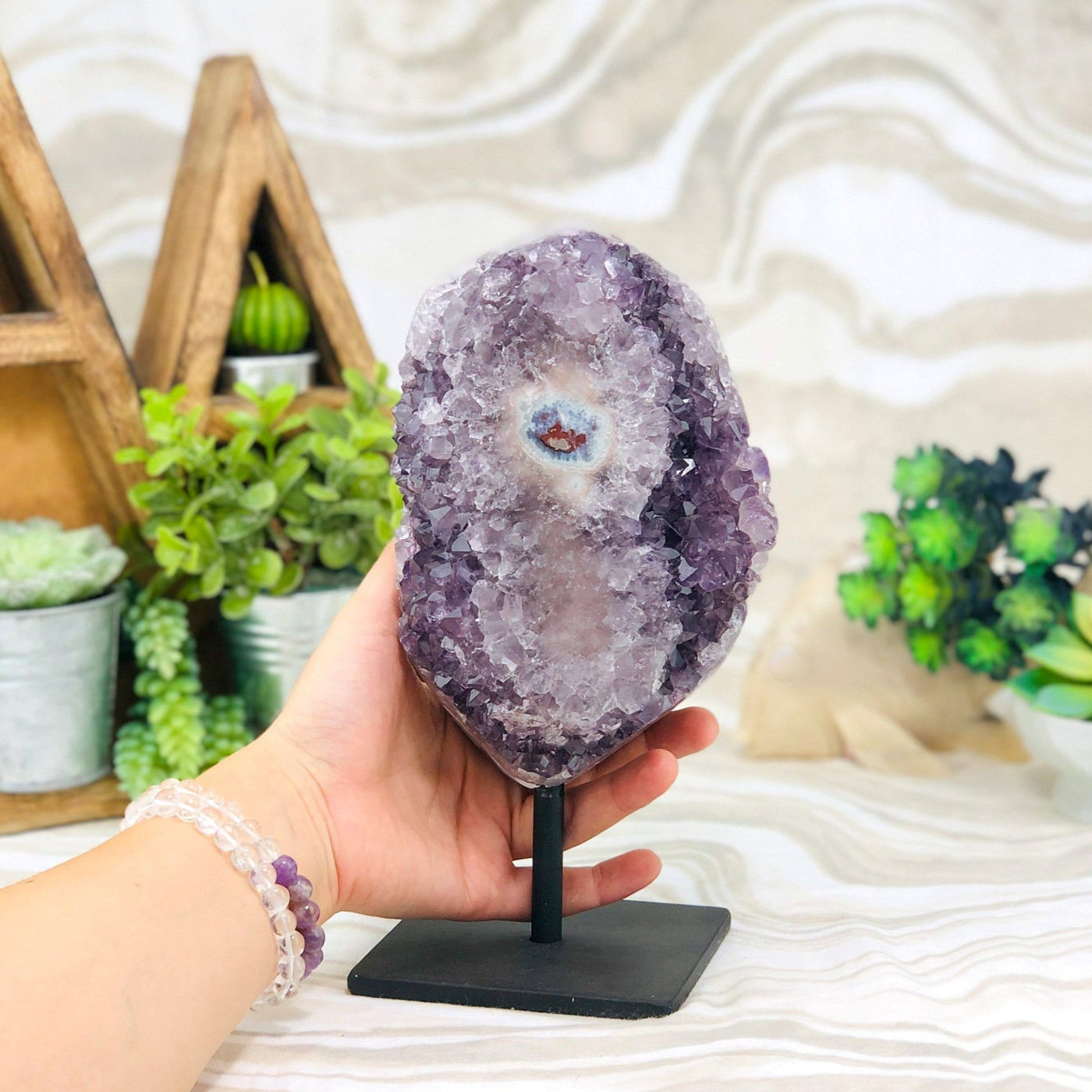 hand hodling up Amethyst Crystal Purple Druzy Semi Polished Stalactite Geode on Stand with decorations in the background