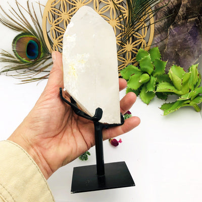 hand holding up Crystal Quartz Point Stone on Metal Stand with decorations in the background