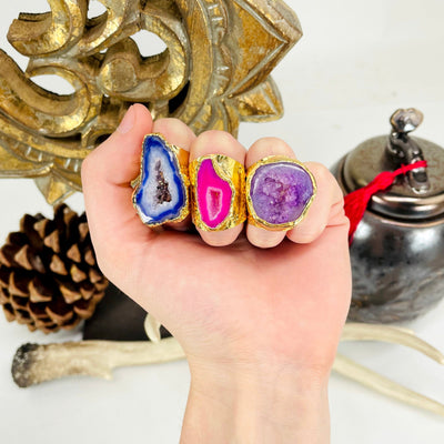 one of each colored agate druzy gold adjustable ring options on hand in front of background decorations for size reference and color comparison