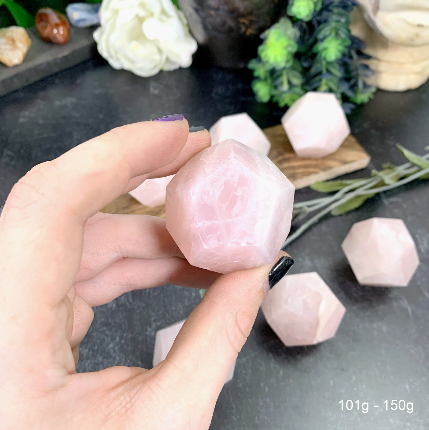 Hand holding up 100-150g Rose Quartz Dodecahedron with others and decorations blurred in the background