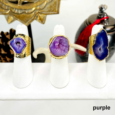 three purple agate druzy gold adjustable rings on ring display in front of background decorations for possible variations 