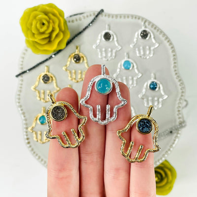 three different hamsa hand pendant options in hand for size reference and possible options with many others in the background