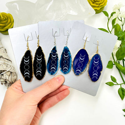 three different colored agate moon phase dangle earrings in their packaging in hand for size reference and possible variations