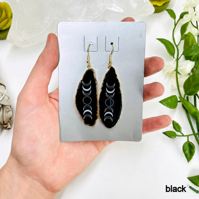 one gold pair of black agate moon phase dangle earrings in their packaging in hand for size and color reference