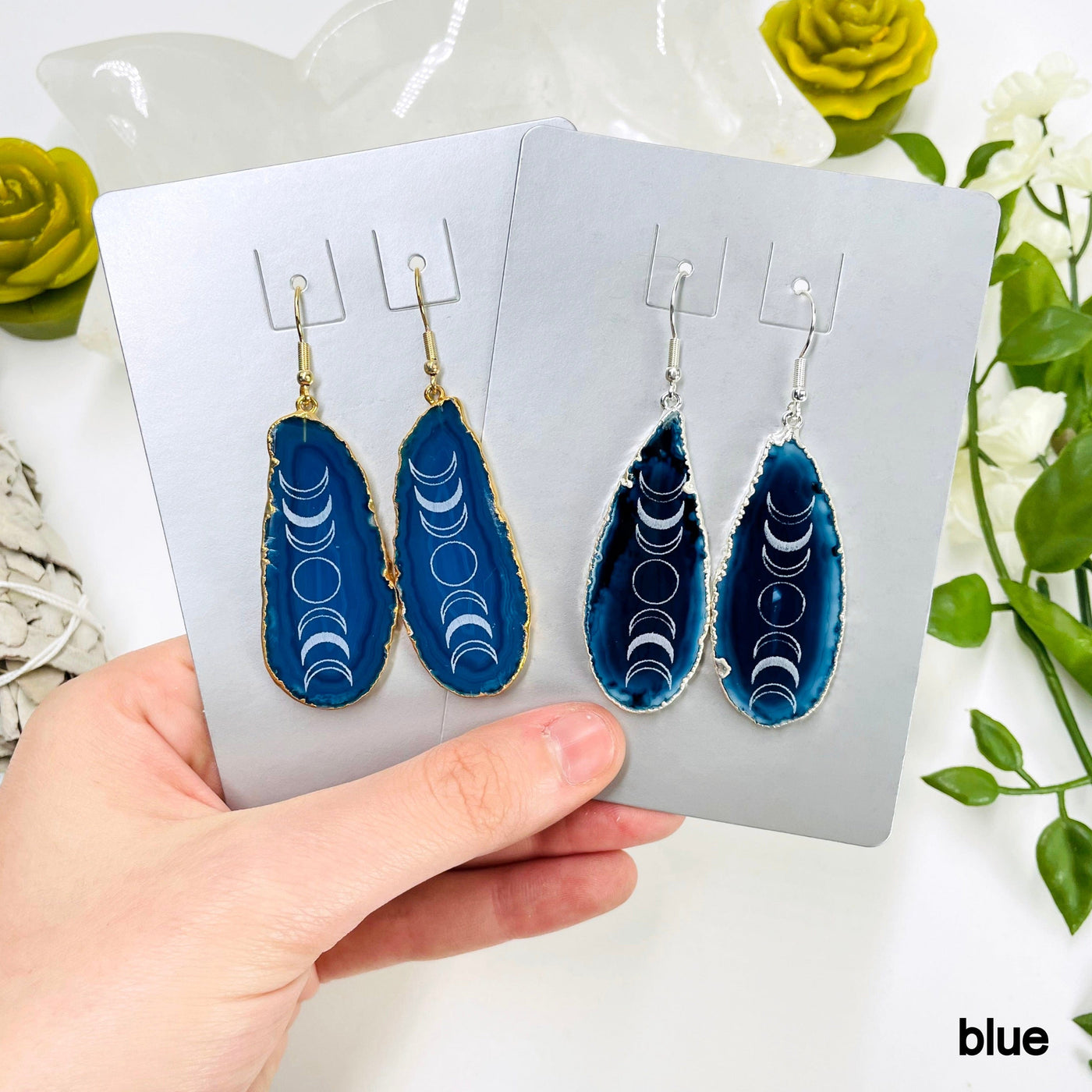 one gold and one silver pair of blue agate moon phase dangle earrings in their packaging in hand for size and color reference