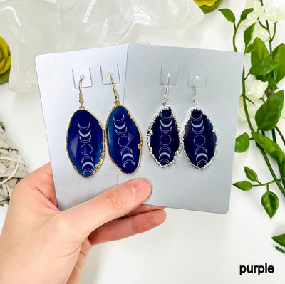 one gold and one silver pair of purple agate moon phase dangle earrings in their packaging in hand for size and color reference