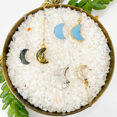 overhead view of one gold and one silver turquoise, labradorite, and crystal quartz crescent moon pendant necklaces in a bowl full of small pebbles for possible options