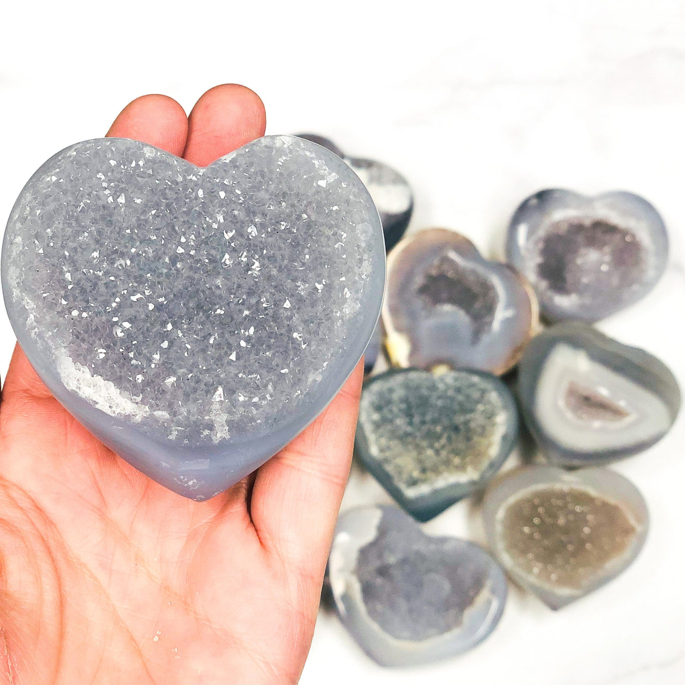 Close up of agate heart with druzy in a hand. Multiple agate hearts in the background on a white surface.
