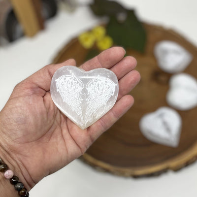 Selenite Stone Heart - Selenite Heart with Angel Wing Engraving in a hand with others in background