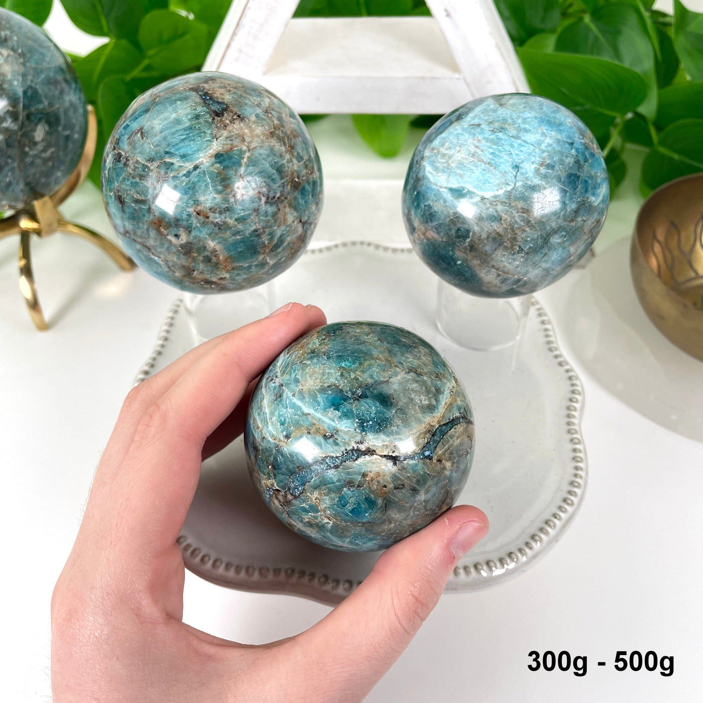 three 300g - 500g blue apatite polished spheres on display in front of backdrop for possible variations with one in hand for size reference