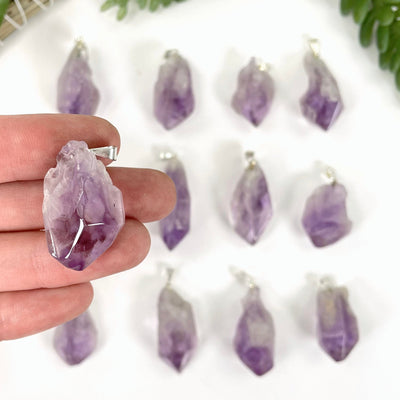 close up of one tumbled amethyst pendant with silver bail in hand for size reference with many others in background