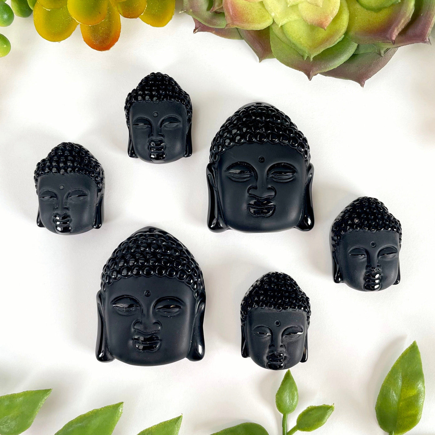 four small and two large black onyx buddha head beads on white background with plant decoration surrounding them for size comparison and possible variations 