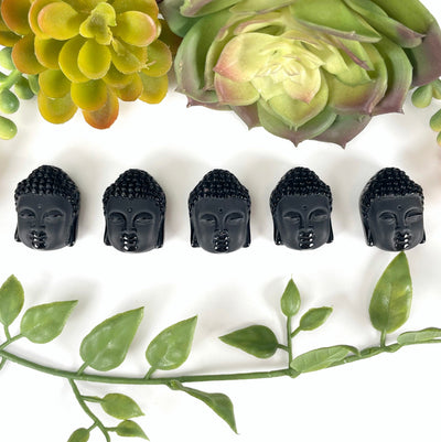 overhead view of five small black onyx buddha head beads on white background with plant decorations for possible variations