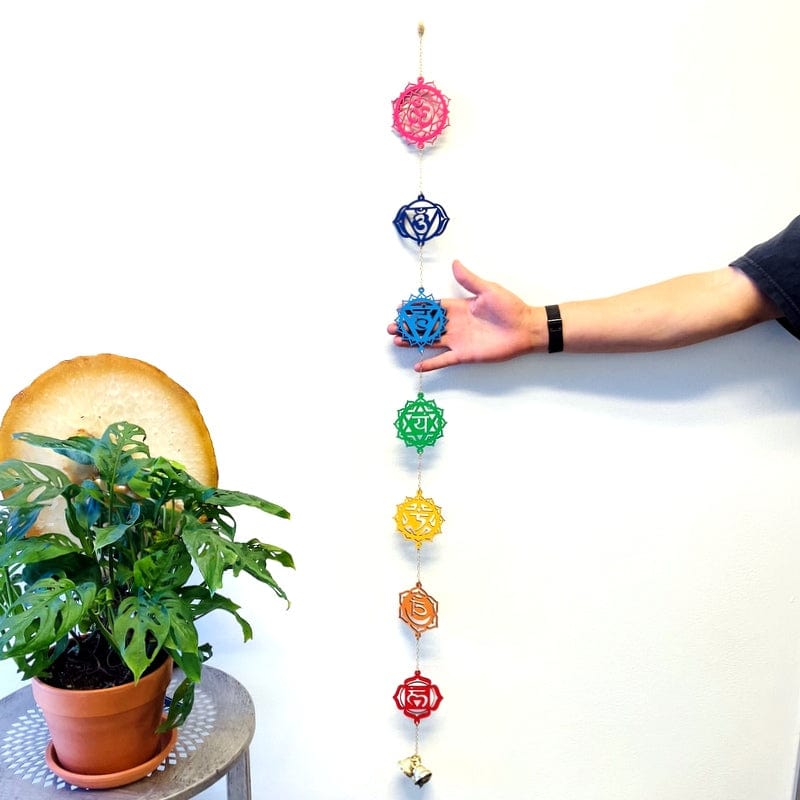 colorful wooden wall hanger with seven chakra symbols and three bells accent on the bottom next to a hand showing the size of the symbols and length of wall hanger 