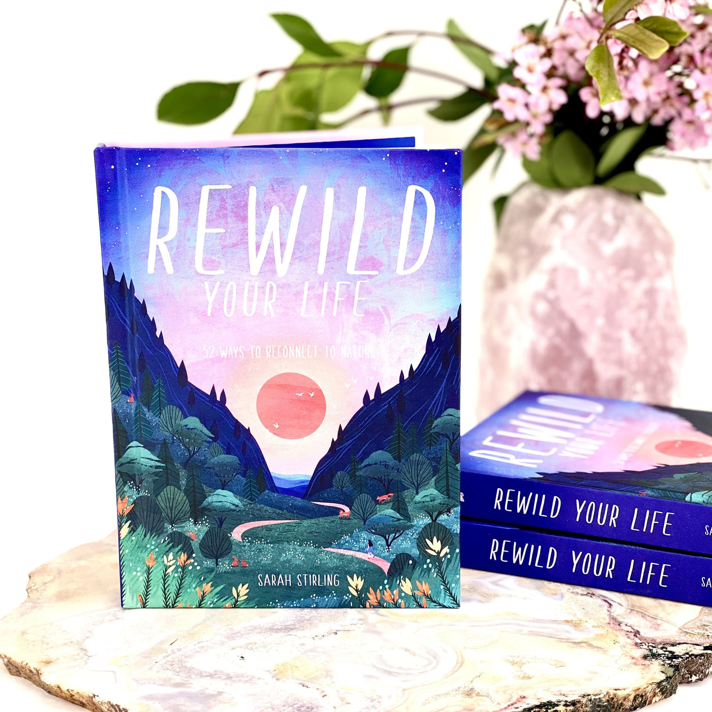 Rewild Your Life Book standing up next to a stack of 2 books with a crystal vase with flowers blurred in the background