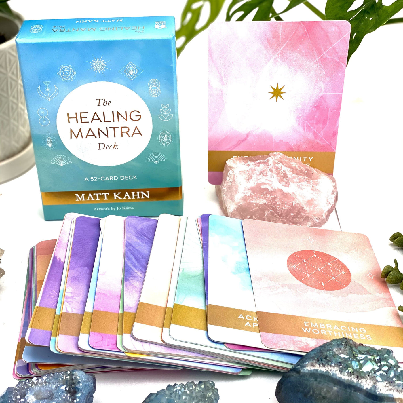 The Healing Mantra Card Deck in teal color box with a pink book and cards on a flat surface to see , colors of cards are in pink teal purple, and green
