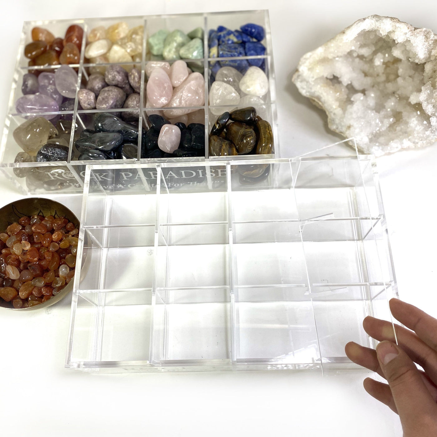 An empty acrylic box shown from the top. A filled crystal box in the background along with half a geode and a bowl of crystals.