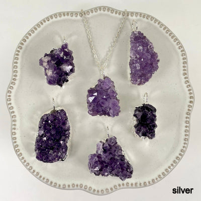 many silver freeform amethyst druzy cluster pendants on display on white background with one of them on a silver chain