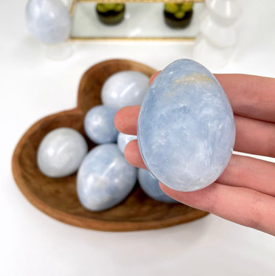 close up of one blue calcite polished egg in hand for size reference and details with many others in a heart bowl in the background