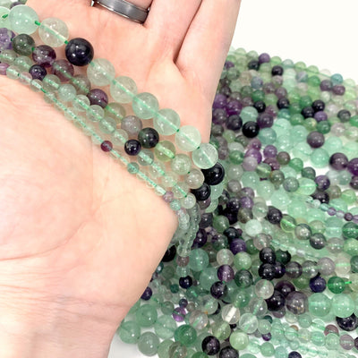 ALL 4 VARIATIONS OF FLUORITE BEADS IN HAND WITH MORE FLUORITE BEADS IN THE BACKGROUND ON A SOLID WHITE BASE BACKGROUND