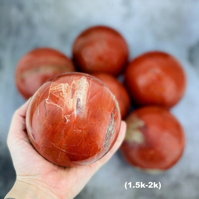 Hand holding up 1.5-2k Red Jasper Sphere with others blurred on gray background