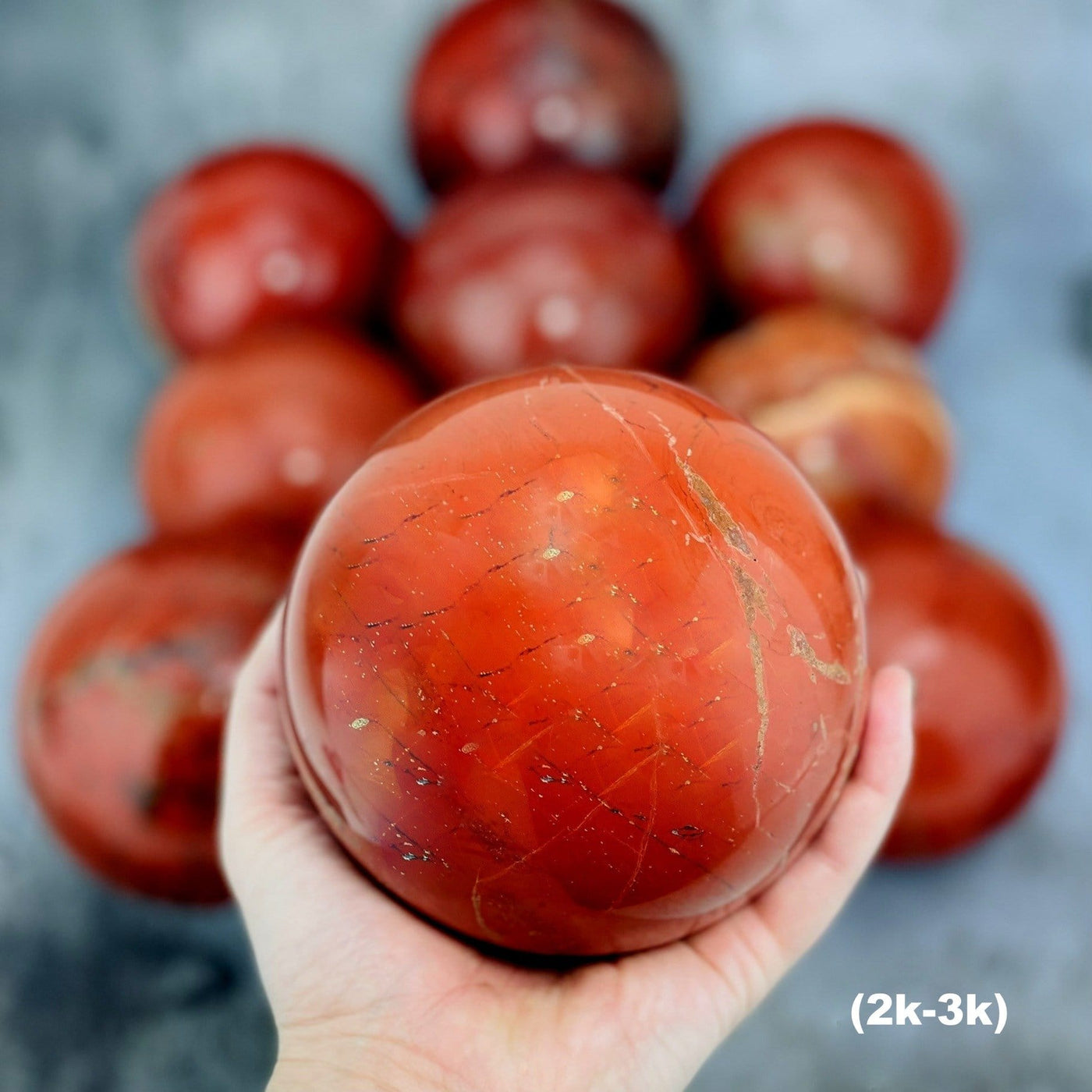 Hand holding up 2-3k Red Jasper Sphere with others blurred on gray background