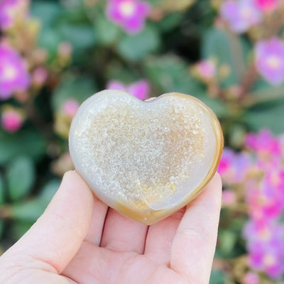 Druzy Natural Agate Heart Shaped Stone front facing in a hand. Flowers are in the background.