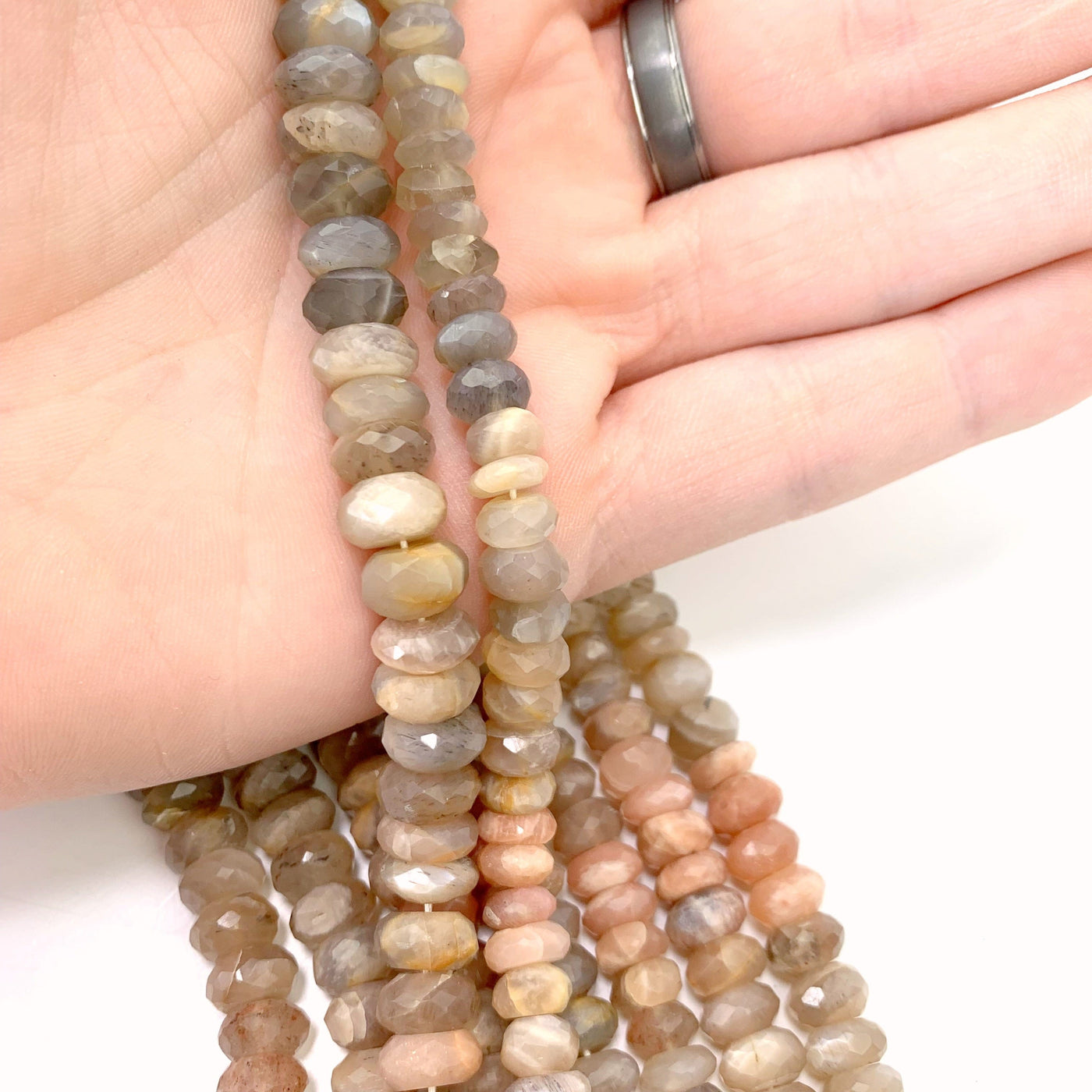 2 moonstone bead strands in hand with more moonstone beads in background wth a white backdrop