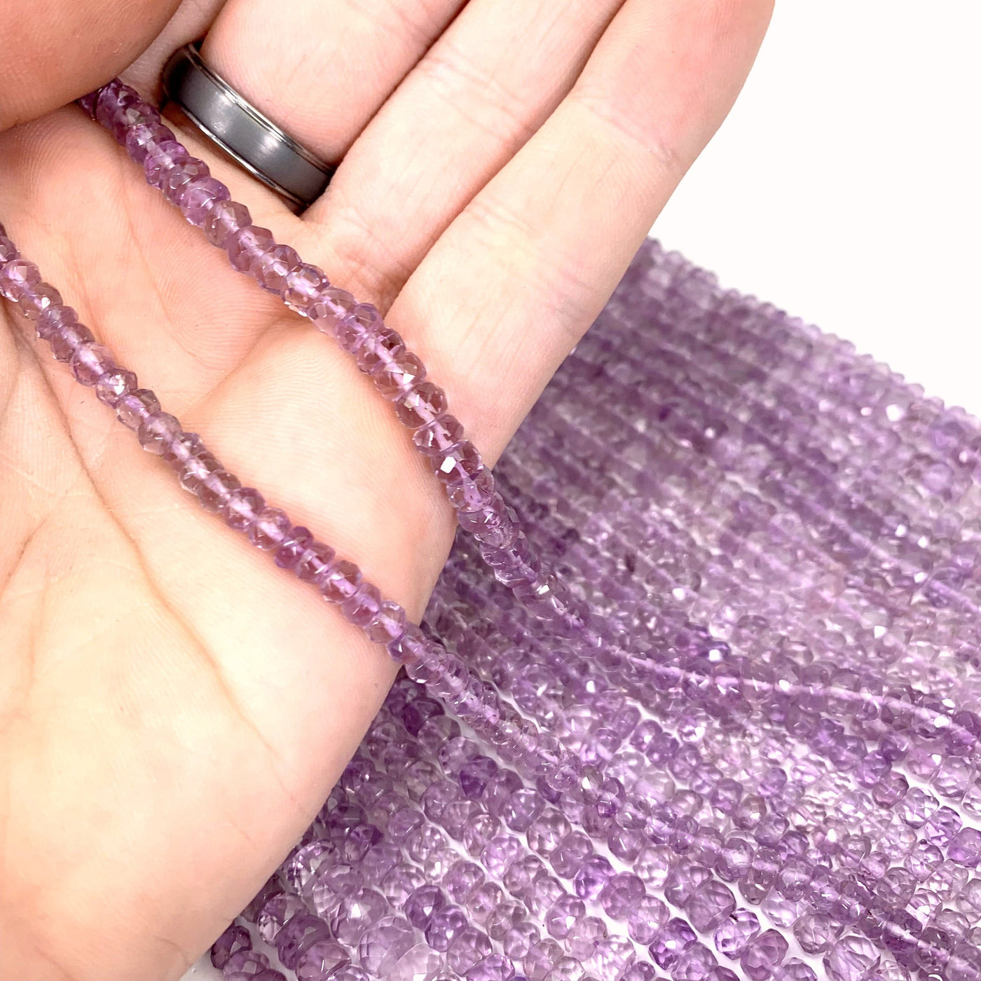 both amethyst bead strands in hand ( left small, right large) with more amethyst beads in the background on a white background