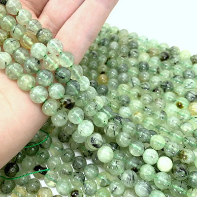 4 strands of prehnite beads in hand with more prehnite beads in the background with a white backdrop