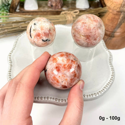 three 0g - 100g sunstone polished spheres on display for possible variations with one in hand for size reference