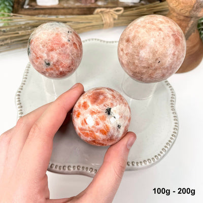 three 100g - 200g sunstone polished spheres on display for possible variations with one in hand for size reference