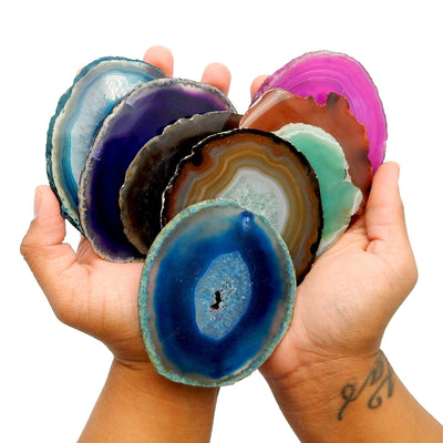 several agate slices in the palm of two hands to show size reference and they come in various colors such as teal purple black natural green red orange pink blue 