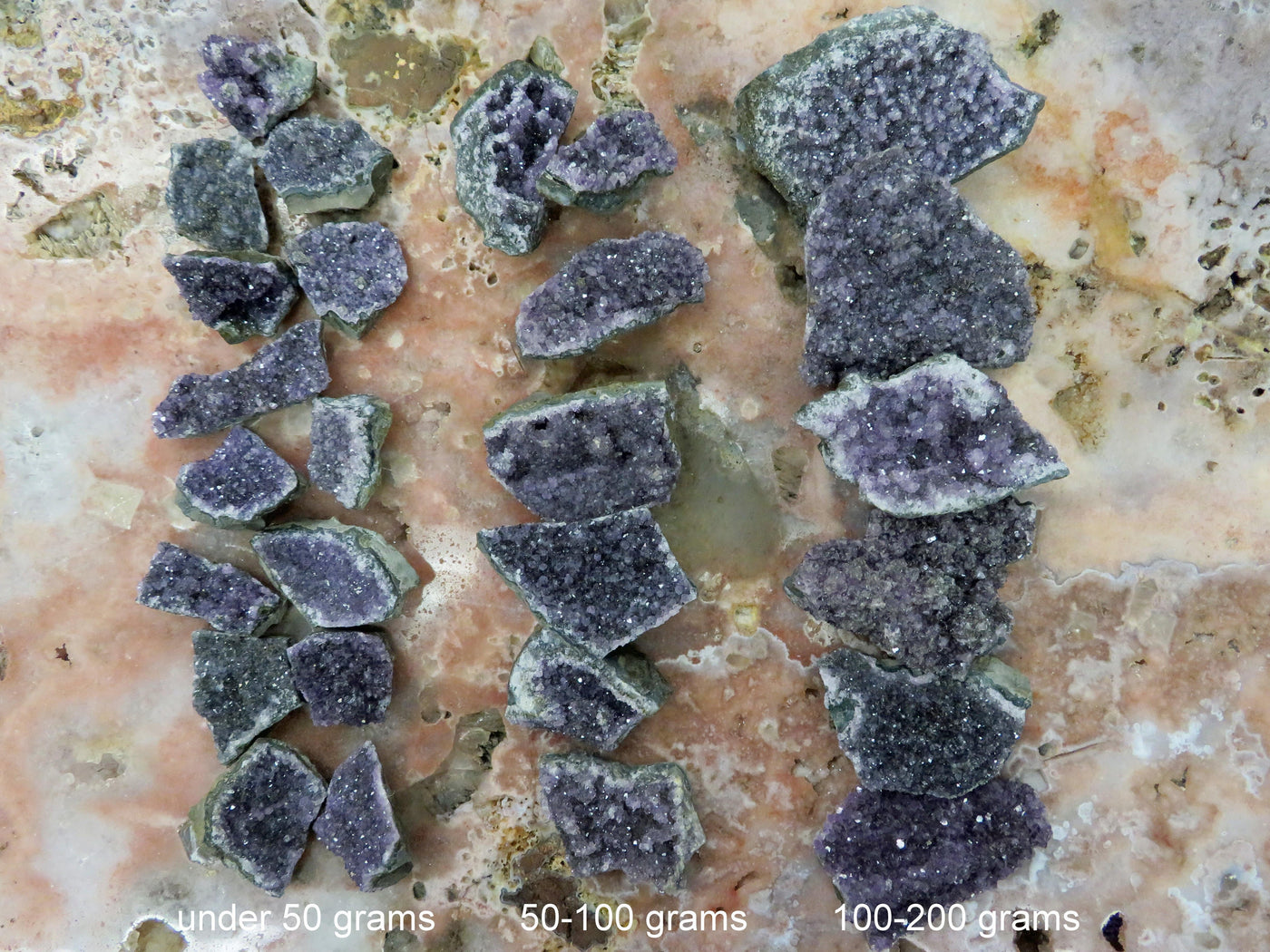 Amethyst clusters are being divided into 3 different weight categories in this picture, all three rows are lined up next to each other. the amethyst clusters are being displayed on a marbled surface.