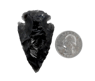 Black obsidian arrowhead next to a quarter showing it is almost twice the size on a white background.