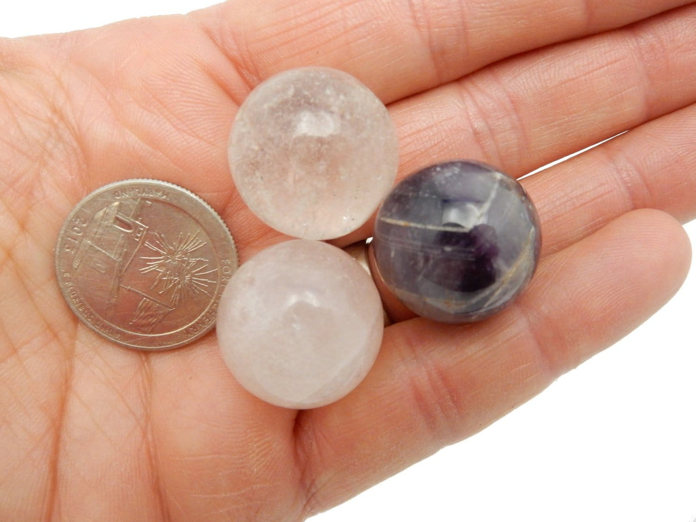 Photo of three spheres one in crystal quartz, one in amethyst, one in rose quartz in a hand with a quarter.  They are about the size of the quarter.